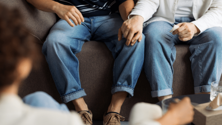 Two people sitting on a sofa hold hands while talking to a therapist.