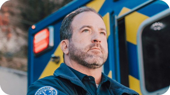 A serious-looking emergency medical worker stands in front of a blue and yellow ambulance, wearing a uniform with an EMS patch on the sleeve.